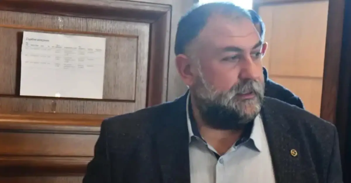 Булфото Мъж с брада и тъмно сако пред дървена стена с информационно табло