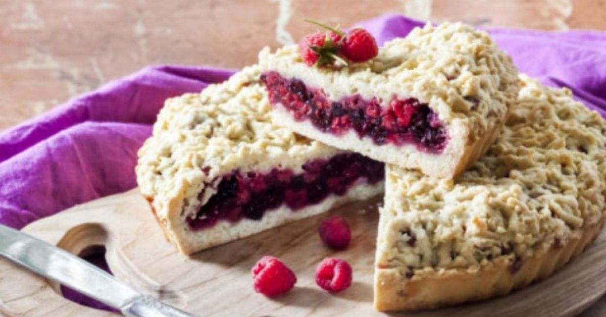 Носталгията на вкуса от детството: настърган сладкиш по бабината рецепта Малинов ронлив пай с пресни плодове, нарязан на дървена дъска.