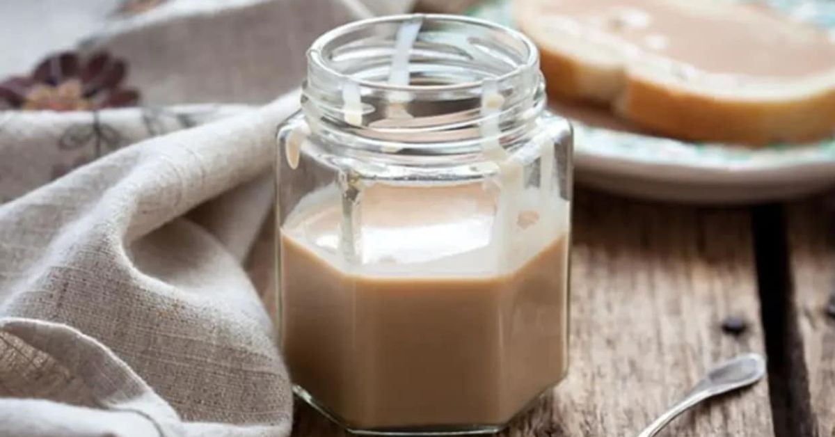 Домашно кондензирано мляко, което е 200 пъти по-вкусно от купешкото Буркан с кремообразен карамелен сос и филия хляб на дървена маса.
