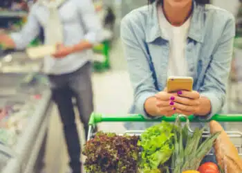 Couple in the supermarket. Cropped image of girl leaning on shopping cart, using a mobile phone and smiling, in the background her boyfriend is choosing food