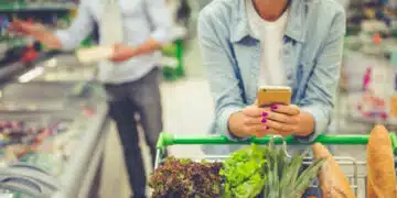 Couple in the supermarket. Cropped image of girl leaning on shopping cart, using a mobile phone and smiling, in the background her boyfriend is choosing food