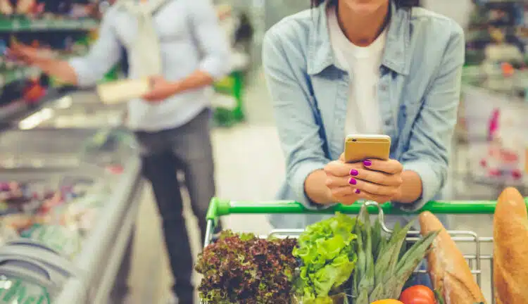 Couple in the supermarket. Cropped image of girl leaning on shopping cart, using a mobile phone and smiling, in the background her boyfriend is choosing food