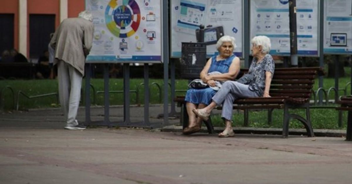 У нас има увеличение на вдовишката пенсия Трима възрастни хора в градски парк, един разглежда плакат, две жени разговарят на пейка.