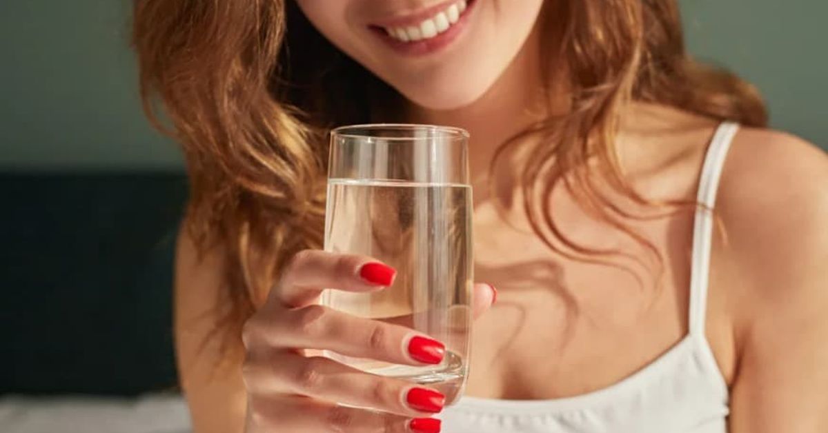 Отговорни за кожата ви: 3 хормона на красотата, които могат да бъдат укротени с храна Усмихната жена с червен маникюр държи чаша вода за хидратация.