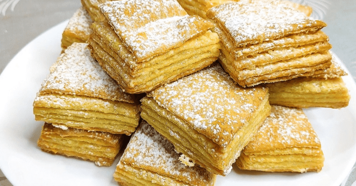 По-вкусно от всяка торта. Бутер тесто за всяко събитие: евтино и изобилно Златни многослойни бутерки, поръсени с пудра захар, на бяла чиния.