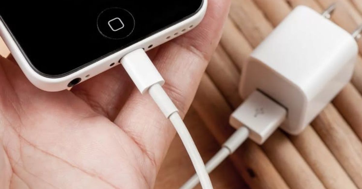 Как да изберете зарядно за смартфон в три стъпки – вземете предвид мощността, кабела и адаптера Човек зарежда бял iPhone с оригинален кабел и зарядно на дървена маса.